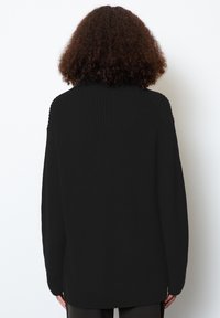Maglione nero a maglia con vestibilità rilassata, pattern testurizzato, dettagli a coste sulle spalle e un retro più lungo, indossato sopra pantaloni scuri.