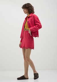 Donna in un completo giacca e gonna a tagli cut-out fucsia brillante sopra un maglione a maglia senape, che indossa scarpe scivolate marrone scuro, in piedi su uno sfondo neutro.