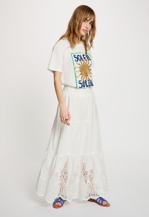 Donna con maglietta bianca con motivo sole e scritta "SOLEIL", gonna lunga bianca con orlo in pizzo e sandali blu, in piedi su sfondo bianco.