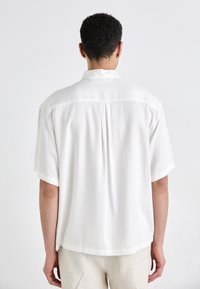 Chemise blanche à manches courtes en tissu léger ; présente un léger éclat, un pli au dos, et l'ourlet tombe en dessous de la taille.