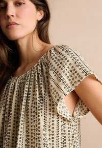 Blouse en crème avec des manches courtes et amples, des motifs floraux noirs verticaux, un col froncé et un détail découpé sur le côté. Tissu léger.