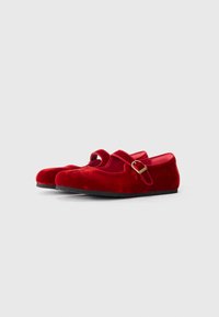 Zapatos Mary Jane de terciopelo rojo con puntera redondeada, una delgada correa en el tobillo y un acento de hebilla dorada. Textura suave con una suela plana negra.