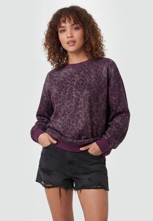 Fiatal nő göndör hajjal, lila leopárdmintás pulóvert és fekete szakadt farmer rövidnadrágot visel, kezei a zsebekben.