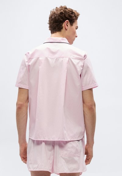Άνδρας που φοράει ροζ ανοιχτό ροζ κοντομάνικο πουκάμισο και σορτς που ταιριάζουν, στέκεται με την πλάτη προς την κάμερα, μπροστά σε ένα απλό λευκό φόντο.