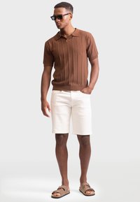 Braun gerippter Strick-Poloshirt mit kurzen Ärmeln, cremefarbene Shorts und braune Slipper; das Modell steht mit entspannter Haltung vor einem grauen Hintergrund.