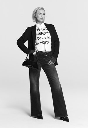 Vrouw met kort blond haar, gekleed in een blazer, een graphic shirt, wijde jeans, zwarte puntige schoenen en met een kleine zwarte handtas in de hand.