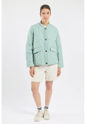 Femme portant une veste matelassée vert clair, un short beige, des chaussettes rayées et des baskets beiges, debout devant un fond clair uni.