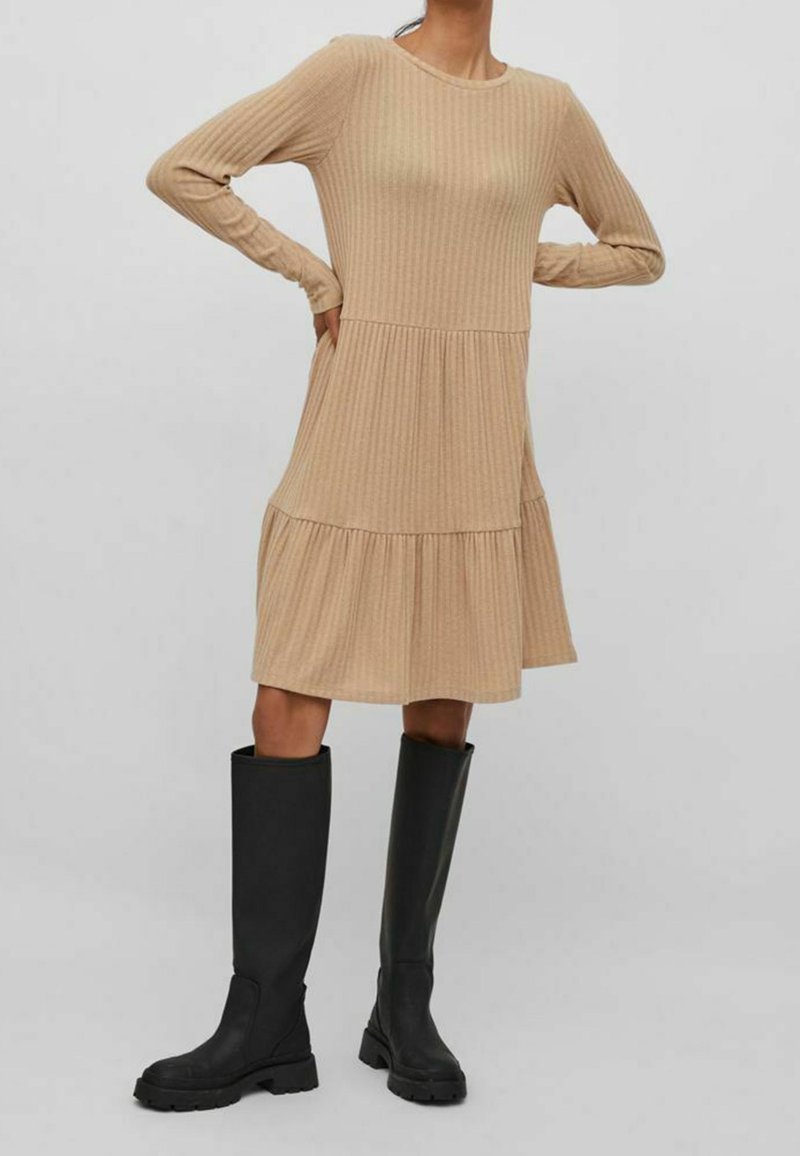 Robe beige côtelée à manches longues avec un ourlet à volants, associée à de longues bottes noires à semelle épaisse.