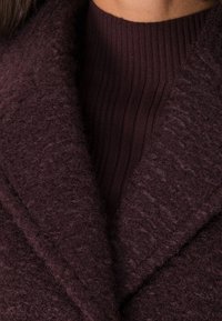 Suéter de punto burdeos con cuello acanalado, que presenta una capa exterior suave y texturada y amplios solapas, creando un efecto de capas.