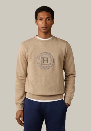 Junger Mann trägt beige Sweatshirt mit rundem Hackett London-Logo und dunkelblaue Hose, steht vor neutralem Hintergrund.