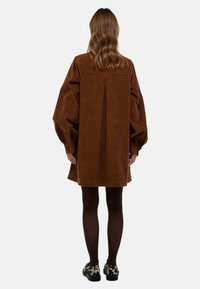 Robe chemise oversize en velours côtelé marron avec des manches bouffantes et un détail de pli dans le dos, associée à des collants foncés et des chaussures à motifs.