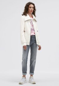 White zip-up hoodie with a wide collar, worn over a pink and white striped shirt. Slim grey jeans and white sneakers with beige accents.