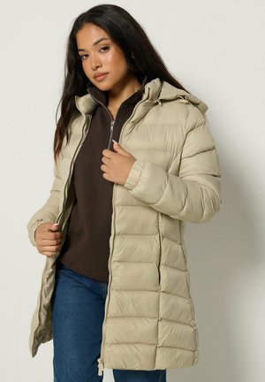 Femme aux longs cheveux foncés portant un manteau matelassé beige jusqu'aux genoux, un haut marron zippé et un jean bleu sur un fond clair uni.