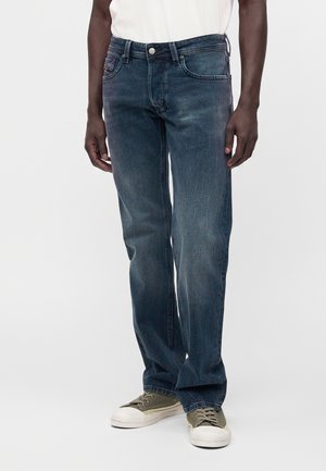 Jean bleu foncé à coupe droite porté par une personne à la peau foncée, associé à des baskets vert olive et blanc et une chemise blanche.