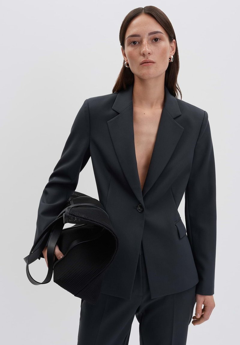 Blazer nero su misura con scollatura a V profonda e chiusura con un bottone. Il modello tiene una borsa a tracolla nera testurizzata con doppi manici.