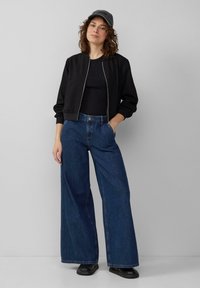 Veste bomber noire, haut noir ajusté, jeans en denim taille haute et large, chaussures noires, et casquette grise avec du texte. Fond clair.