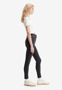 Fiatal nő fehér rövidujjú felsőben, fekete skinny farmerben, fehér zokniban és bézs sportcipőben áll oldalt egy fehér háttér előtt.