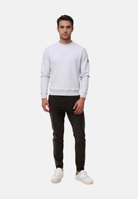 Light gray sweatshirt with ribbed cuffs and hem, featuring a subtle logo on the front. Paired with dark slim-fit pants and white sneakers.