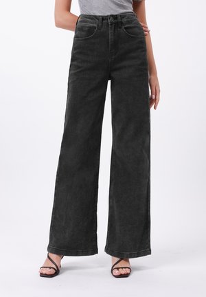 Jean large noir en denim, avec une taille haute, un design classique à cinq poches et un ourlet effiloché. Porté avec des sandales noires à lanières.
