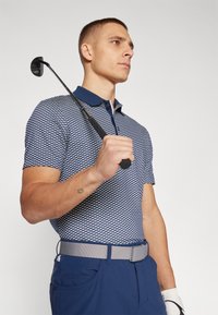 Marinblå och vit mönstrad pikétröja med krage, i kombination med blå byxor och ett grått bälte. Personen håller en golfklubba.