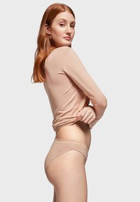 Nude tanga od mješavine pamuka s glatkom završnom obradom, s niskim strukom i nježnim picot rubom. Model nosi dolčevitu u boji kože s dugim rukavima.