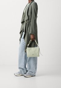 Grüne Ledertasche mit geflochtenem Griff, rechteckiger Form und Reißverschluss. Kombiniert mit einem langen olivgrünen Trenchcoat und hellblauen Jeans.