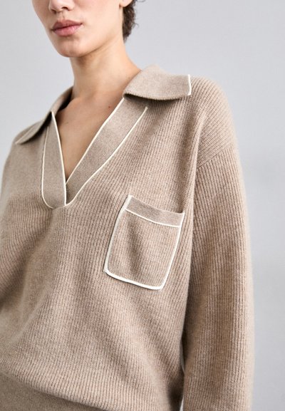 Beige kootud jumpsuit kraega, V-kaelusega ja taskuga. Erineb kontrastse valge äärisega krae ja tasku servadel. Pehme tekstuur.