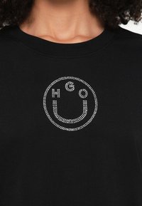 Sweatshirt preta com um design de rosto sorridente redondo e cravejado. Apresenta o texto "HGO" em strass brancos dentro do contorno do sorriso.
