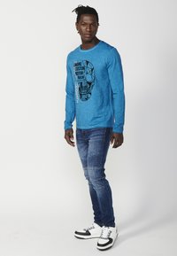 Blue long-sleeve shirt with a black graphic skull design, paired with blue jeans featuring stitched details and white sneakers with black accents.