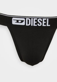 Czarny string wykonany z miękkiego materiału z szerokim elastycznym pasem oznaczonym napisem "DIESEL" na biało. Gładka tekstura i minimalne pokrycie.