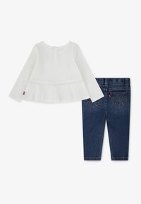 Weißes Langarmoberteil mit Peplumsaum und Knopfverschluss hinten, kombiniert mit dunkelblauen, engen Jeans mit Gesäßtaschen.