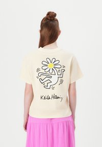 Short-sleeve, cream cotton T-shirt featuring a black and yellow daisy graphic and signature on the back, worn with a pink skirt.