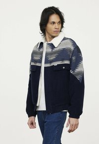 Cordjacke mit marineblauem Grund, die ein Tribal-Muster in Grau und Hellblau aufweist, sowie einen weichen weißen Fleecekragen. Zwei Fronttaschen.