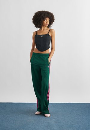 Γυναίκα με κατσαρά μαλλιά στέκεται με τα χέρια στις τσέπες, φορώντας μαύρο crop top Adidas και πράσινο παντελόνι με ροζ ρίγες σε μπλε πάτωμα.