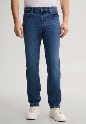 Mann, der blaue Jeans und weiße Sneakers trägt, steht vor einem einfarbigen Hintergrund.