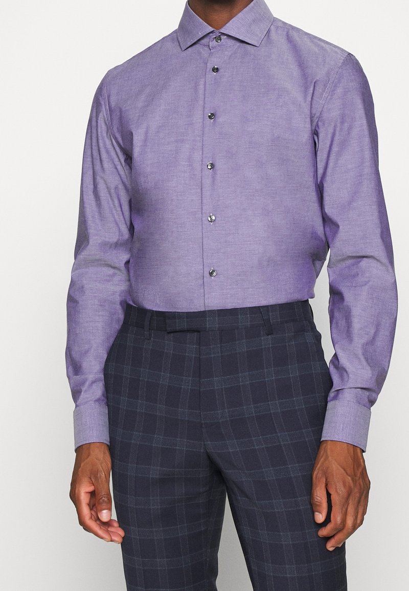Lichtpaars overhemd met knopen en een klassieke kraag, voorzien van zilveren knopen. Draagt bij donkere geruite broek, met een gladde stoftextuur.