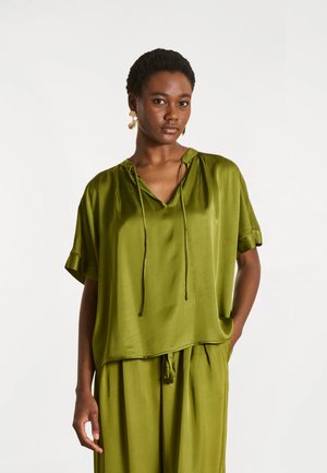 Jeune femme portant un chemisier ample et brillant vert olive à manches courtes et un pantalon assorti, debout avec une main dans la poche sur un fond uni.