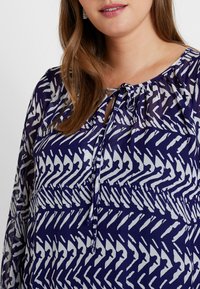 Mørkeblå og hvid mønstret bluse med rund halsudskæring, bindedetalje og lange gennemsigtige ærmer. Stoffet har en let, flydende tekstur.