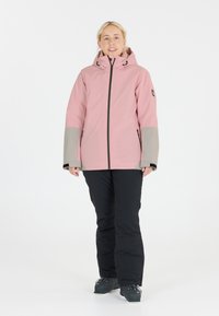 Rosa wasserdichte Jacke mit grauen Ärmeln, Reißverschluss, Kapuze und Seitentaschen; kombiniert mit schwarzen Skihosen und schwarzen Stiefeln. Minimalistisches Design.