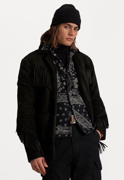 Giacca in suede nera con frange su una camicia nera a fantasia, abbinata a un berretto lavorato a maglia nero. Dettagli notevoli includono tessuti testurizzati e motivi a contrasto.