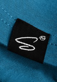 Turquoise stof met een gestructureerde weef heeft een zwart merketiket met een witte gestileerde logo. Stiksel zichtbaar op de achtergrond.