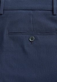 Tissu bleu foncé texturé avec une poche fendue horizontale et un seul bouton à quatre trous sous l'ouverture de la poche.