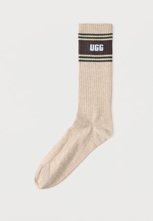 Smėlio spalvos ribuotos kojinės su tamsiai ruda juosta viršuje, ant kurios yra balti "UGG" užrašai ir plonos žaliai baltos juostelės virš ir po juosta.