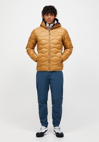 Mann iført en sennepsbrun quiltet hettegenserjakke, blå bukser og hvite sneakers, står med hendene i lommene på jakken.