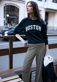 Sudadera negra con estampado blanco "BOSTON 1980", combinada con pantalones anchos beige texturizados. La modelo sostiene una chaqueta negra con forro blanco.