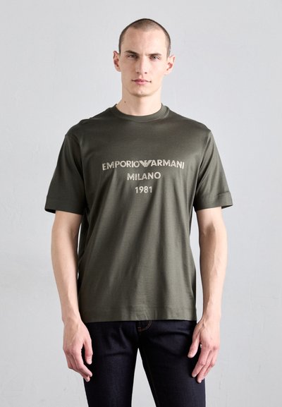 Oliwkowa, krótka koszulka z okrągłym dekoltem. Na przodzie nadruk "EMPORIO ARMANI MILANO 1981" w kolorze białym. Wykonana z gładkiego materiału.