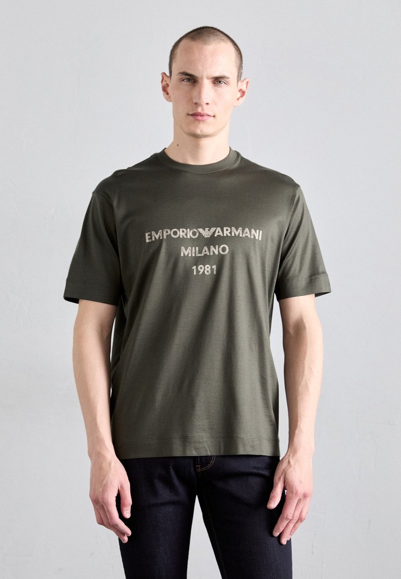 Olive roheline lühikeste varrukatega t-särk ümmarguse kaelusega. Esiküljel on valge kiri "EMPORIO ARMANI MILANO 1981". Valmistatud siledast kangast.