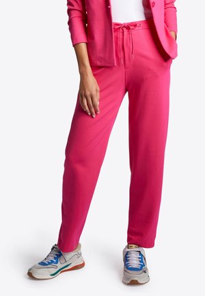 Vrouw die felroze joggingsbroek en blazer draagt met witte en blauwe sneakers, staat met één hand in de zak en de andere ontspannen.