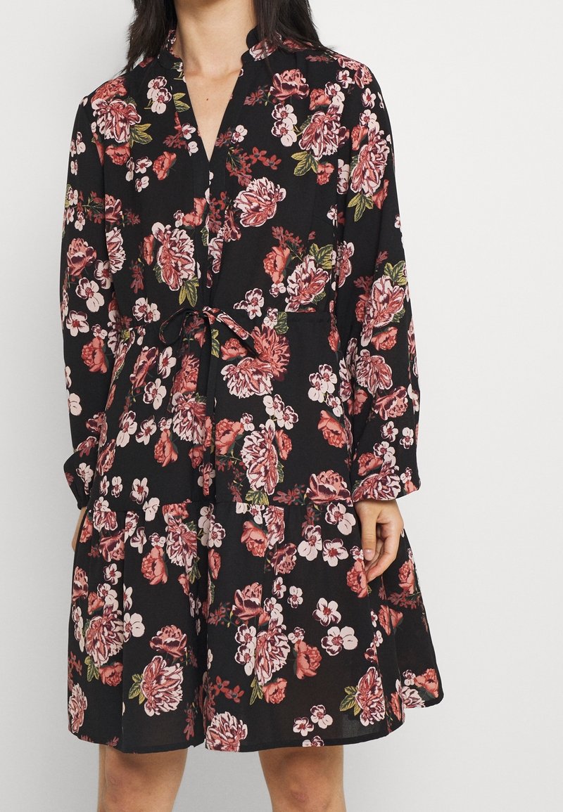 Černé květinové šaty s dlouhými rukávy, stahováním v pase a sukni ve vrstveném provedení. Tkanina má strukturovaný povrch s růžovými a bílými květinovými vzory.