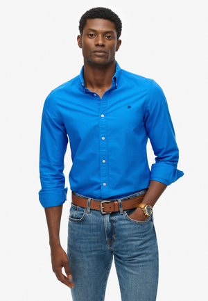 Blauw bloesje, van katoen, lange mouwen opgerold, klein logo op de borst, gecombineerd met een bruine riem en blauwe spijkerbroek.
